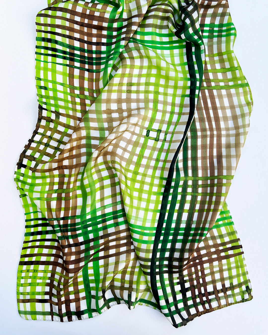 Aflangt silketørklæde i grønne farver, silketørklæde i brune farver, Silketørklæde lyst ternet, Silketørklæde med grønne og brune håndmalede tern, Silketørklæde aflangt 160 x 65 cm, Silketørklæde i 100 % ren silke, silketørklæde i ægte silke twill, silketørklæde med design af Susanne Schjerning, silketørklæde med rullede kanter. Giv et silketørklæde i gave. Bæredygtigt silketørklæde i bæredygtig dansk design, silketørklædet der er nemt at binde. 